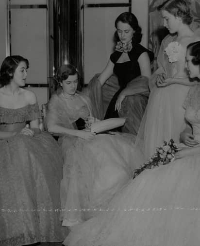 Five women in formal gowns gathered indoors, two seated, three standing, appearing to read or discuss a document.