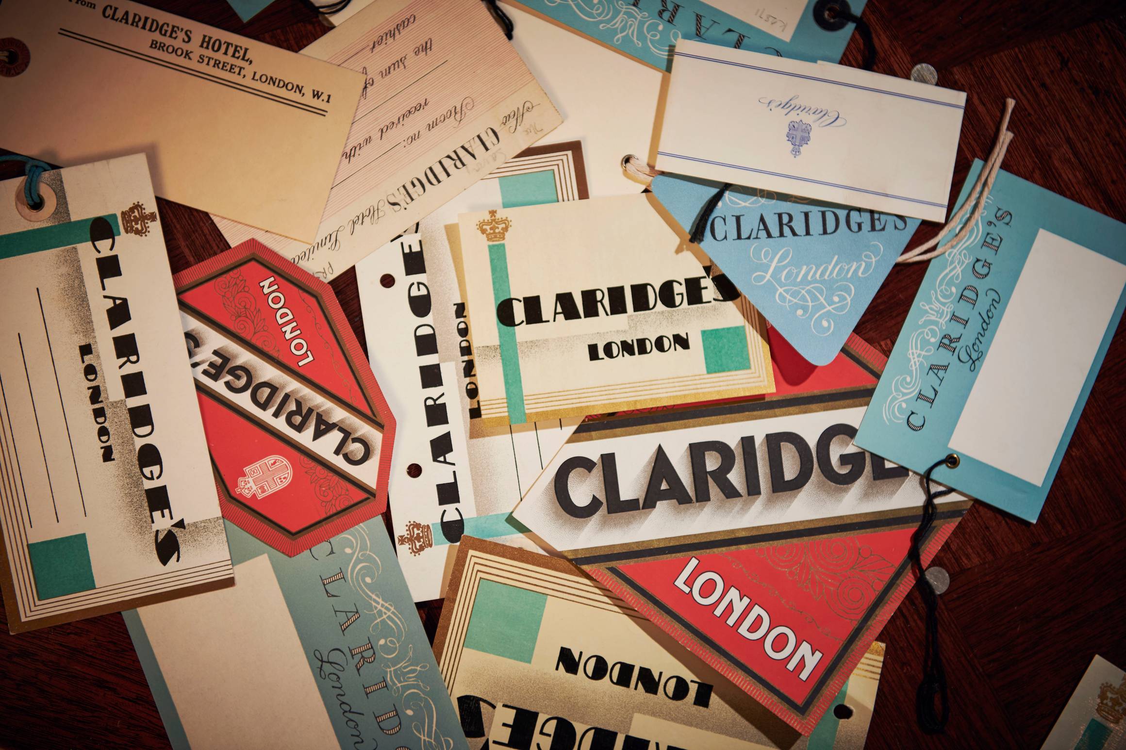 Vintage Claridge’s luggage tags in assorted colours and styles, scattered on a dark wooden surface.