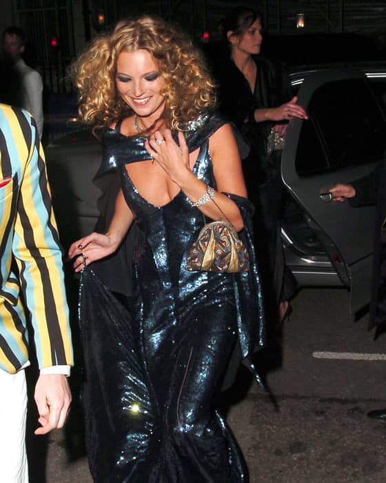 Kate Moss in sparkling blue evening gown stepping out of a car, smiling, holding dress and patterned handbag.