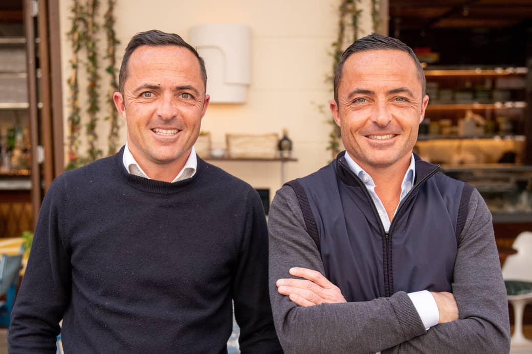 Two individuals standing side by side in a casual outdoor setting, both dressed in smart yet relaxed attire, smiling warmly towards the camera with a backdrop of greenery and a stylish café interior.