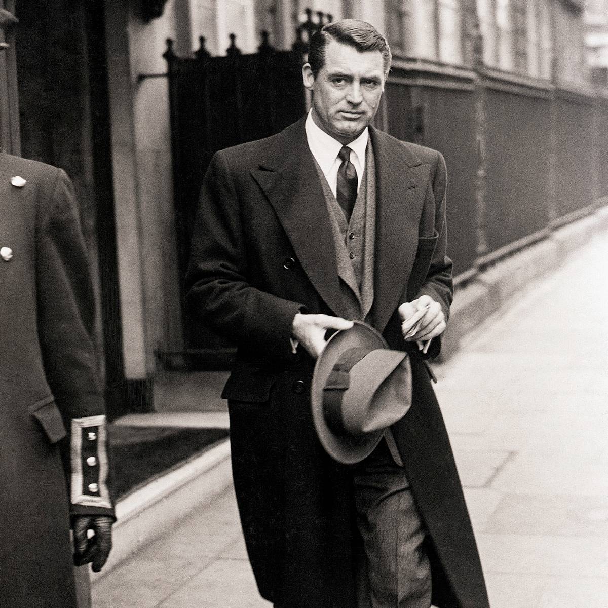 Cary Grant in overcoat and suit walking on city sidewalk, holding a fedora, with a uniformed doorman partially visible.