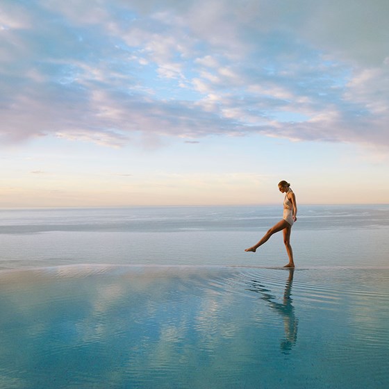 A lady in a swimsuit walks along the infinity pool edge infront of the sea and a beautiful sunset